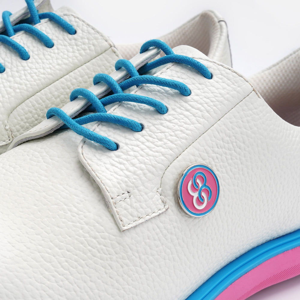 White sneaker with blue laces and a pink and blue logo on a white background sells at Paradise Golf